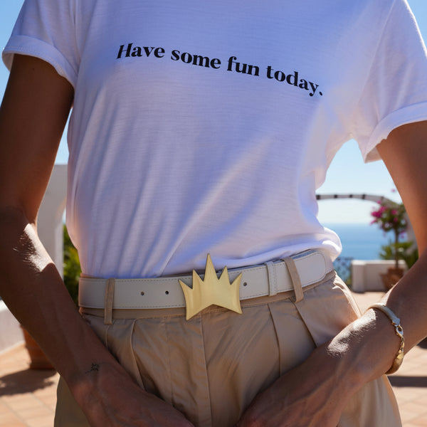 A person wearing a white graphic T-shirt with the text 'Have some fun today.' in black, paired with beige trousers and a beige belt featuring a gold crown buckle, photographed outdoors with a blurred scenic background.