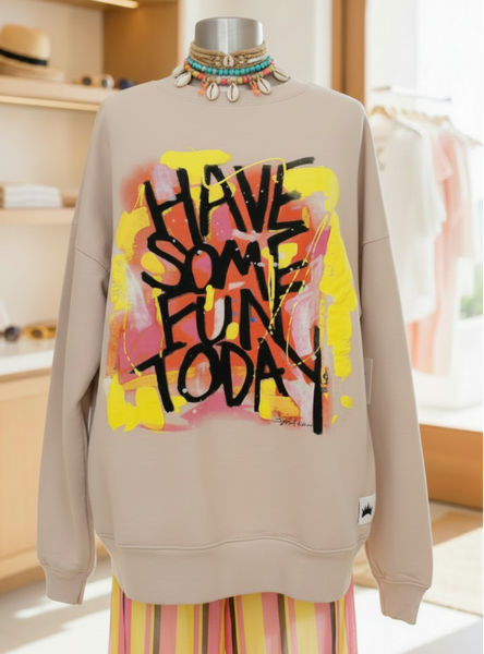 A beige crewneck sweatshirt displayed on a mannequin torso in a bright boutique interior. The sweatshirt features a large graphic print on the front with a yellow and pink paint-splatter background and the text 'HAVE SOME FUN TODAY' in black handwritten lettering.