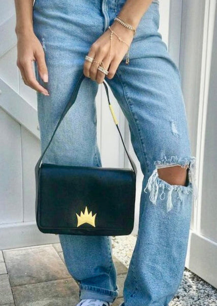 A person wearing light blue ripped jeans holds a black shoulder bag with a gold crown logo. The person is also wearing gold bracelets and rings, standing on a stone tile floor next to a white door.