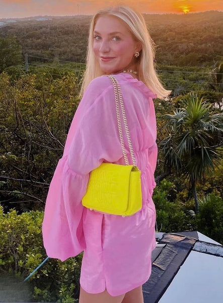 Woman in pink outfit with yellow bag standing in a scenic outdoor setting at sunset.
