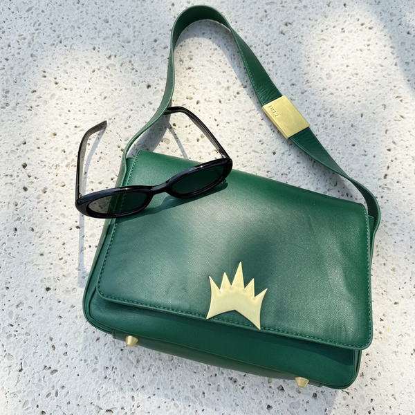 Green handbag with a gold crown design and sunglasses on a stone surface