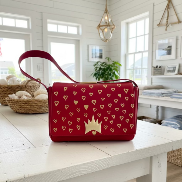 A red handbag with a gold crown and heart pattern, displayed on a white wooden table in a bright, modern room.