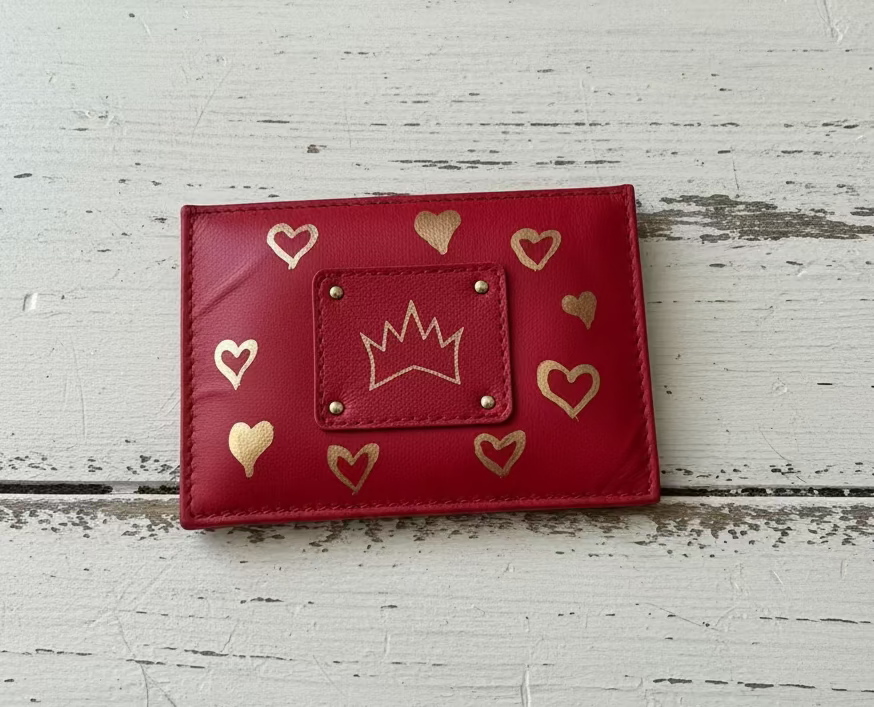 A red faux leather wallet with gold heart and crown details, displayed on a rustic white-painted wooden surface.