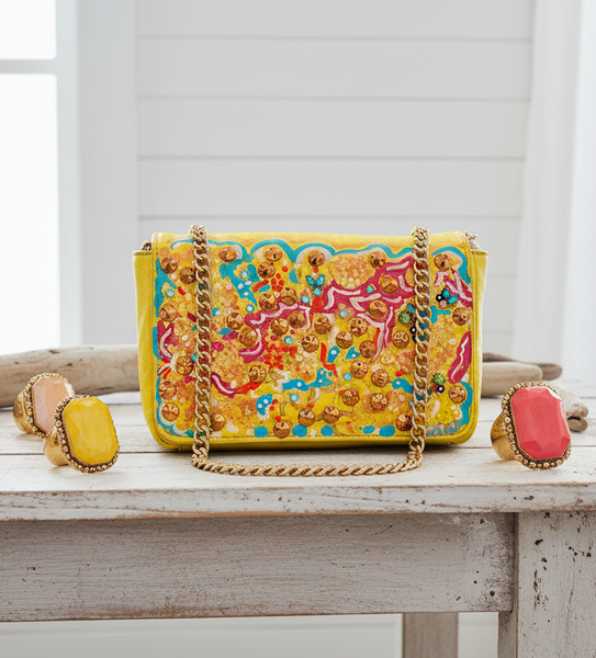 A bright yellow embroidered handbag with a gold chain strap sits on a rustic white table, surrounded by colorful statement rings in a bright, airy room setting.