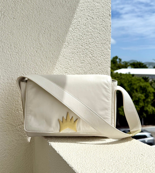 A white lambskin shoulder bag with custom crown logo hardware and a brushed gold finish on the strap, displayed against an outdoor background.