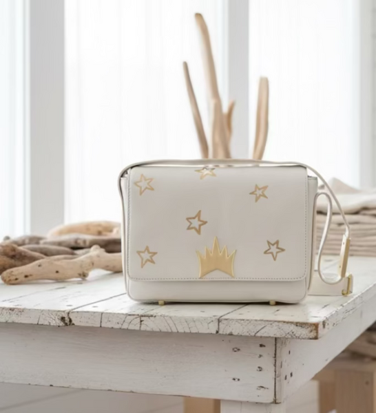 A cream-colored leather cosmetic pouch with gold star and crown motifs sits on a rustic white wooden table in a bright, airy boutique setting.