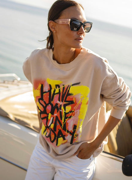 A woman wearing an oversized beige crewneck sweatshirt with vibrant text-based artwork in yellow, red, and black on the front.