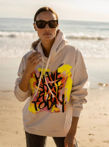A person wearing an oversized beige hoodie with colorful text-based artwork on the front, standing on a beach.