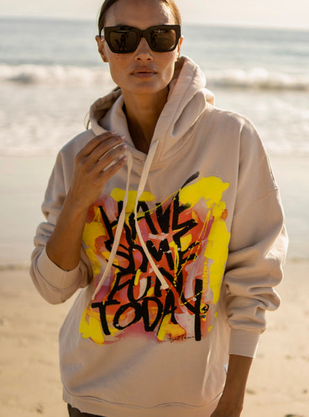 A person wearing an oversized beige hoodie with colorful text-based artwork on the front, standing on a beach.