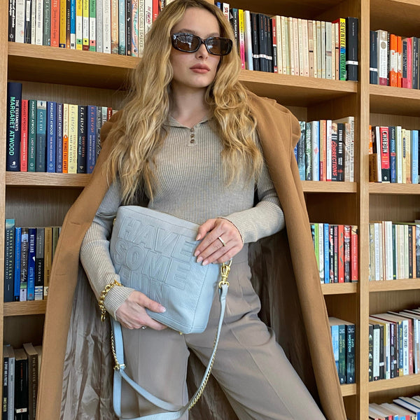 A woman sitting in front of a bookshelf wearing sunglasses, a grey pebbled cow leather bag with lamb trim, and a beige coat.