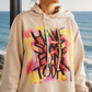 A woman wearing a beige hoodie with a colorful graffiti-style graphic on the front, standing in front of a large window with a view of the ocean.