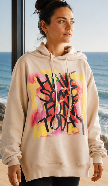 A woman wearing a beige hoodie with a colorful graffiti-style graphic on the front, standing in front of a large window with a view of the ocean.