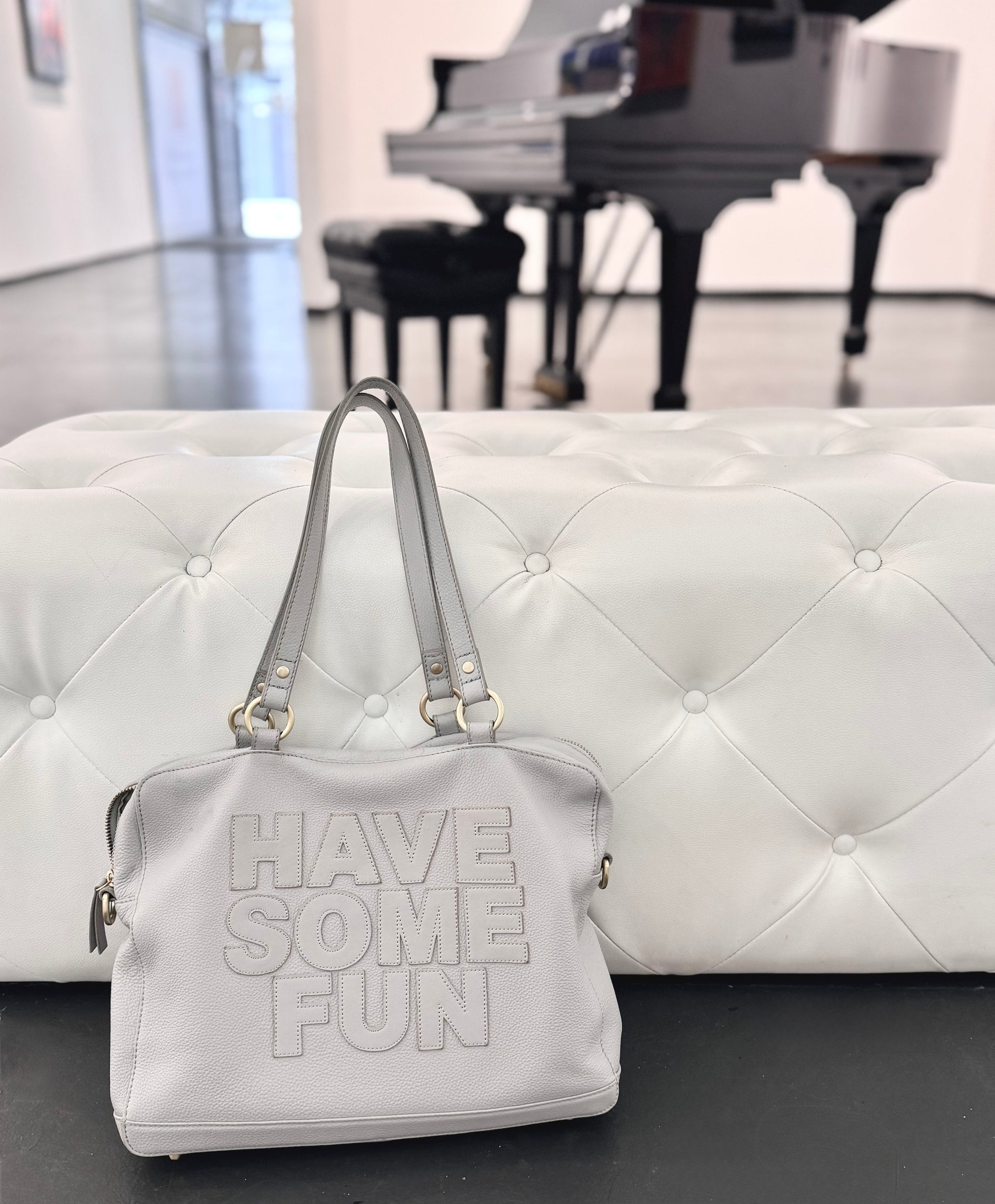 A light gray faux-leather handbag with the words 'HAVE SOME FUN' embossed on the front, resting on a white tufted bench in front of a blurred piano in a gallery space.