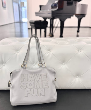 A light gray faux-leather handbag with the words 'HAVE SOME FUN' embossed on the front, resting on a white tufted bench in front of a blurred piano in a gallery space.