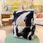 A hand-painted canvas backpack with black and white brushstrokes, featuring a small gold crown emblem on the front flap and a white top handle, displayed on a mint green table in a modern living room setting.