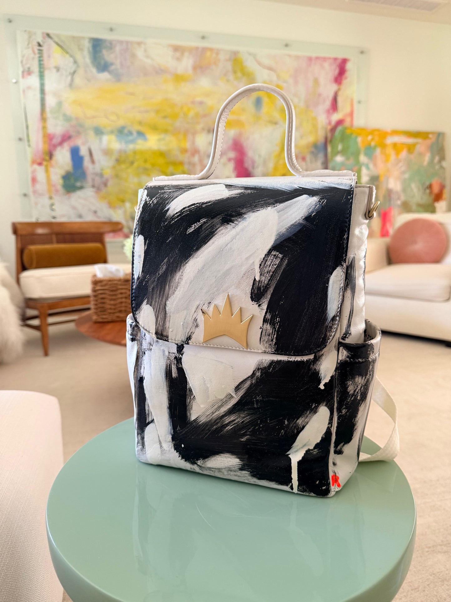 A hand-painted canvas backpack with black and white brushstrokes, featuring a small gold crown emblem on the front flap and a white top handle, displayed on a mint green table in a modern living room setting.
