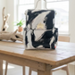 A hand-painted canvas backpack with a black and white abstract brushstroke pattern, featuring a gold crown emblem on the front flap and a top handle, resting on a light-colored sofa.
