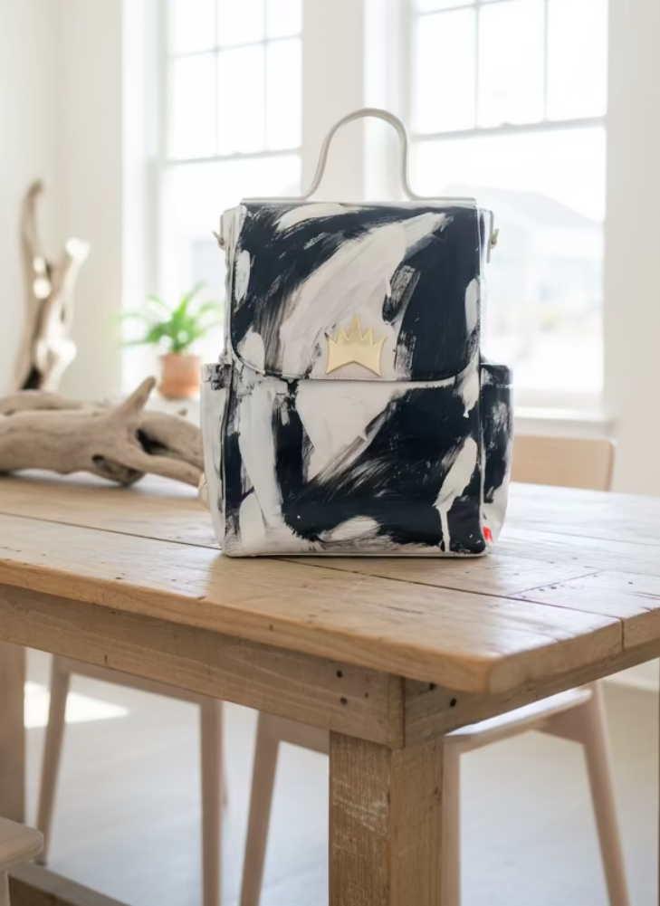 A hand-painted canvas backpack with a black and white abstract brushstroke pattern, featuring a gold crown emblem on the front flap and a top handle, resting on a light-colored sofa.