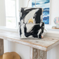 A hand-painted canvas backpack featuring a black and white abstract brushstroke pattern with a small gold crown emblem on the front pocket. The backpack has a white top handle and is displayed on a textured beige rug in front of a dark wooden panel.