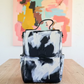 A black and white abstractly painted canvas backpack with a top handle, shown in a home interior setting.
