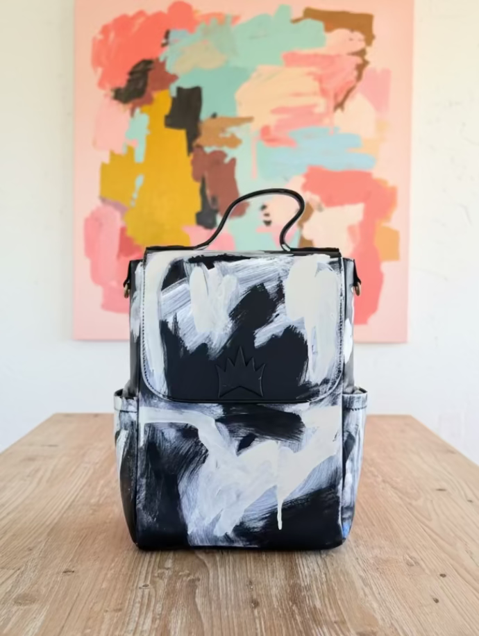 A black and white abstractly painted canvas backpack with a top handle, shown in a home interior setting.