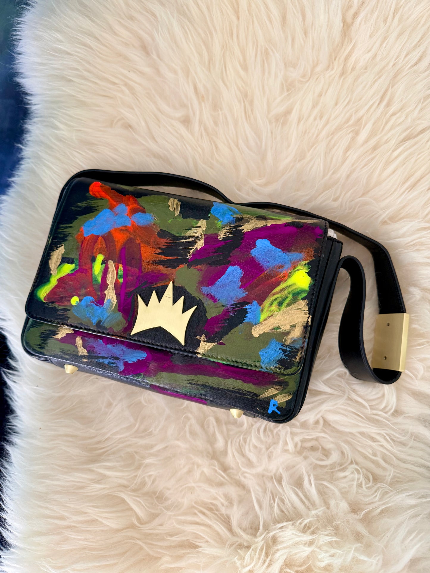 A rectangular shoulder bag with a glossy black frame and strap, featuring a vibrant multicolor abstract paint-splatter design on the front flap. The bag has a prominent gold crown logo on the center of the flap and is displayed on a white faux fur surface.
